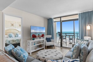A modern living room with a large TV, a round coffee table, and a balcony with a view of the ocean. - Oceanfront Beach Cove w/ Resort Pools-Wide Balcony (North Myrtle Beach)
