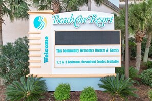 Een bord voor Beach Cove Resort, dat eigenaren en gasten verwelkomt en aangeeft dat er appartementen met 1, 2 en 3 slaapkamers aan het strand beschikbaar zijn.