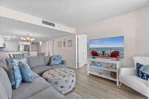 A modern living room with a grey sofa, a round ottoman, and a white armchair. There is a flat-screen TV mounted on the wall, a dining area in the background, and a view of the ocean through the window. - Oceanfront Beach Cove w/ Resort Pools-Wide Balcony (North Myrtle Beach)