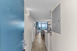 A hallway leading to a balcony with a view of the sea, a kitchen area with a dining table, and a white door. - Oceanfront Beach Cove w/ Resort Pools-Wide Balcony (North Myrtle Beach)