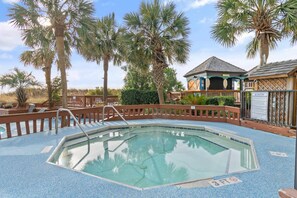 An outdoor swimming pool with a wooden deck, surrounded by palm trees and a building with a tiled roof. - Oceanfront Beach Cove w/ Resort Pools-Wide Balcony (North Myrtle Beach)