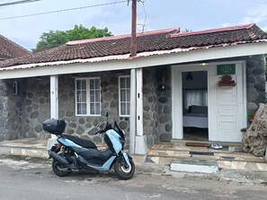 Un bâtiment en pierre avec un toit en tuiles, une moto garée devant, et une porte qui donne sur une pièce contenant un lit et une petite table.