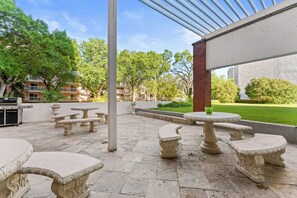 An outdoor patio area with stone tables and benches, a grill, and a view of a building and trees.