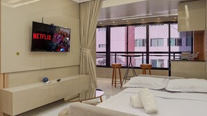 Habitación de hotel moderna con una televisión grande mostrando Netflix, una cama con ropa blanca, una mesita con taburetes y vistas a un edificio rosa a través de ventanales amplios.
