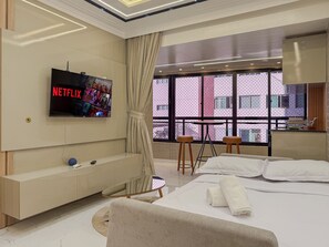 Ein modernes Hotelzimmer mit großem Fernseher, der Netflix zeigt, einem Bett mit weißen Leinen, einem kleinen Tisch mit Hocker und Blick auf ein rosafarbenes Gebäude durch große Fenster.