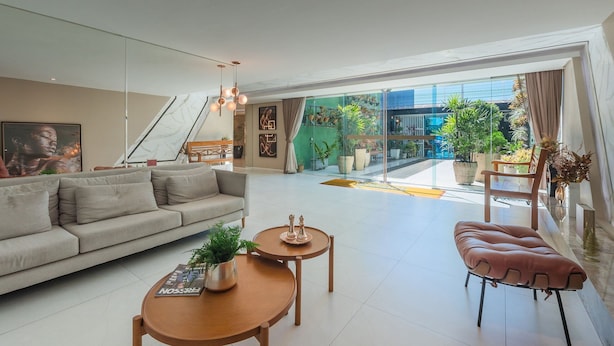 Una sala de estar moderna con un sofá, una mesa de centro y un otoman, un comedor con una mesa y sillas, y grandes ventanales que ofrecen vistas al exterior.