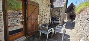 A stone building with a wooden door, a table with chairs, and a grill outside. - Auvergne country house "La maison du Cantou (Vieillevie)