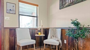 Ein Zimmer mit zwei Stühlen, einem kleinen Tisch mit Kerze, einem Fenster mit Jalousien und einem an der Wand hängenden Gemälde.