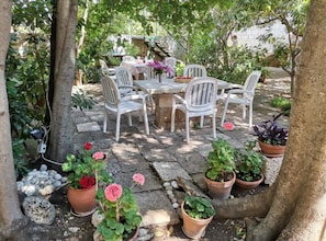 Un patio al aire libre con una mesa de piedra y sillas blancas, rodeado de macetas con plantas y árboles.