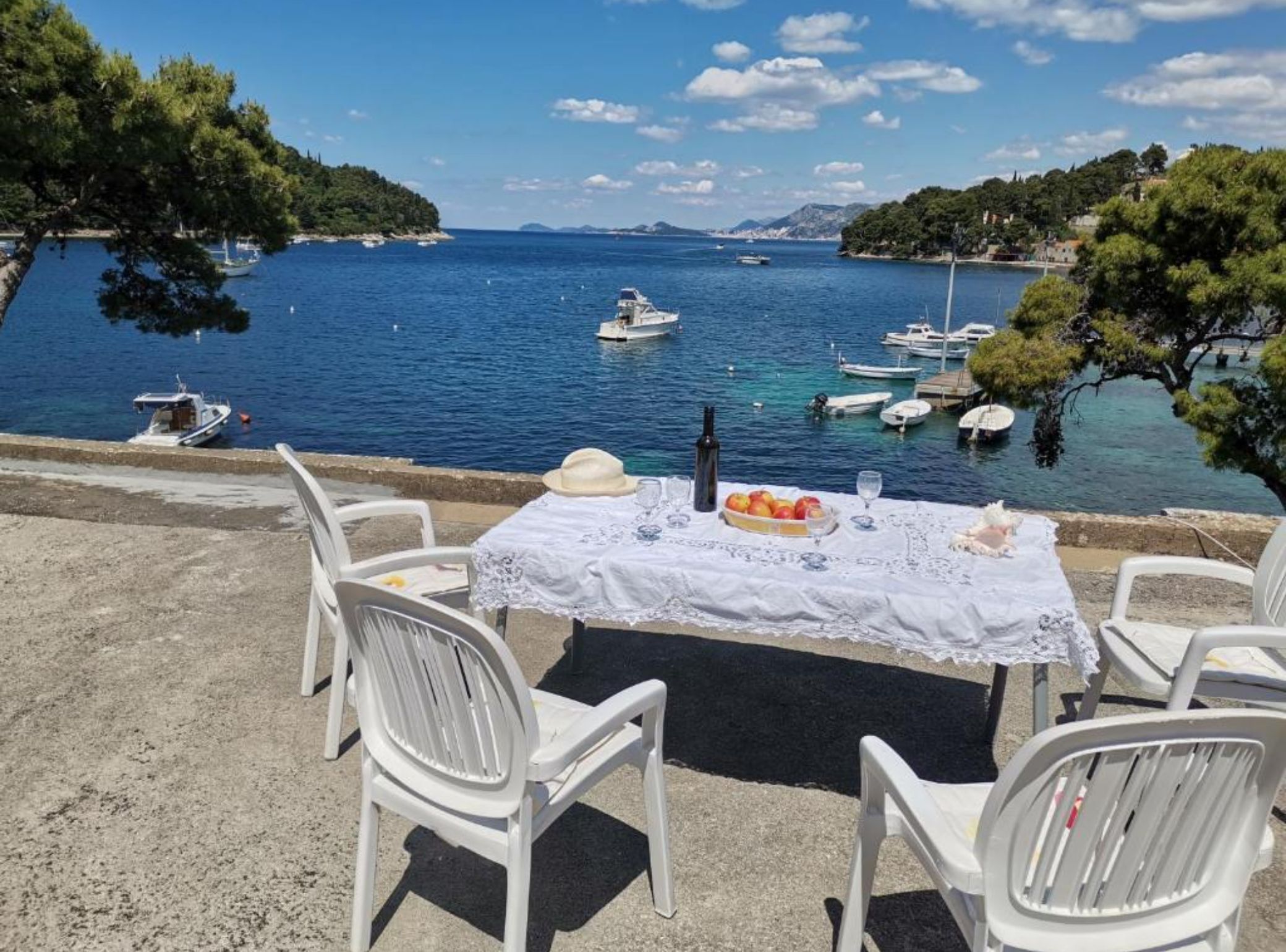 A table set with a bottle, a hat, and fruit on a tablecloth, overlooking a marina with boats.