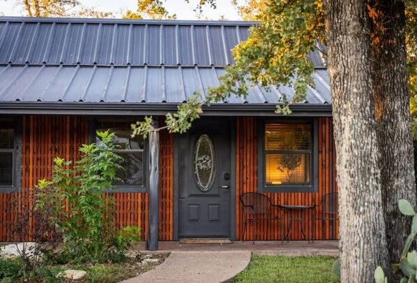 A building with a dark metal roof, wooden paneling, a black door with a decorative glass panel, and a small outdoor seating area.