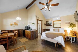 A room with a bed, a desk, a kitchenette, a ceiling fan, and a door leading to another room.