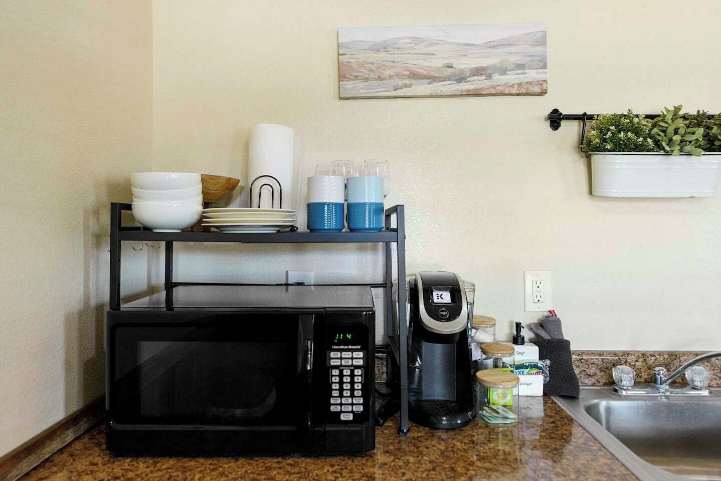 A kitchen with a microwave, coffee maker, and a shelf with dishes and cups.