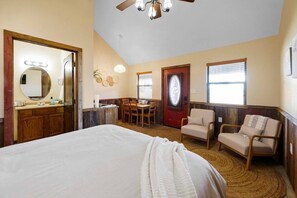 A bedroom with a large bed, a desk, two chairs, a door, and a ceiling fan.
