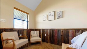 A room with a wooden paneled wall, a window with blinds, two chairs, a woven rug, and three framed pictures on the wall. - The Fantail at Nicole Creek Cabins (Waco)