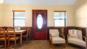 A dining area with a wooden table and chairs, a red door, and two windows with blinds. - The Fantail at Nicole Creek Cabins (Waco)