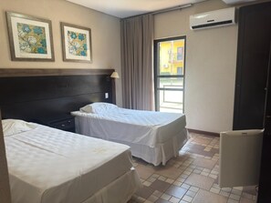 A hotel room with two beds, a headboard, a window with curtains, an air conditioning unit, and a refrigerator.