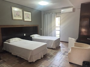 A hotel room with two beds, a chair, an air conditioning unit, and a window with curtains.