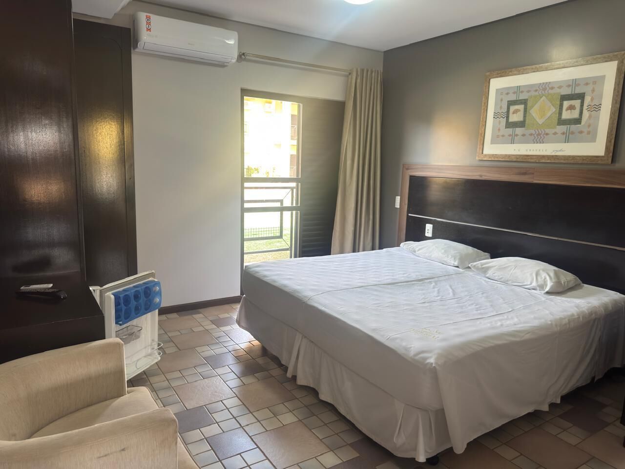 A hotel room with a bed, a chair, an air conditioning unit, a window with curtains, and a framed picture on the wall.