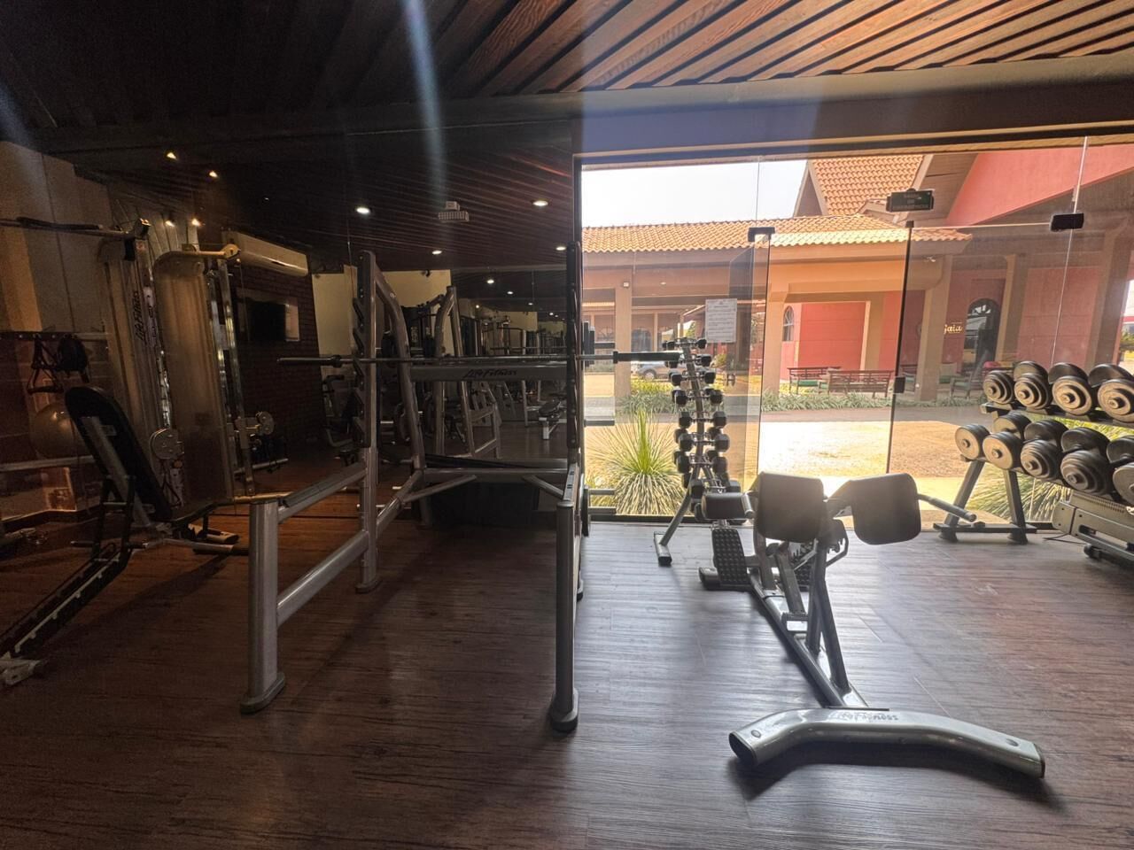 A gym with various exercise equipment, a wooden floor, and a view of the outside through a glass door.