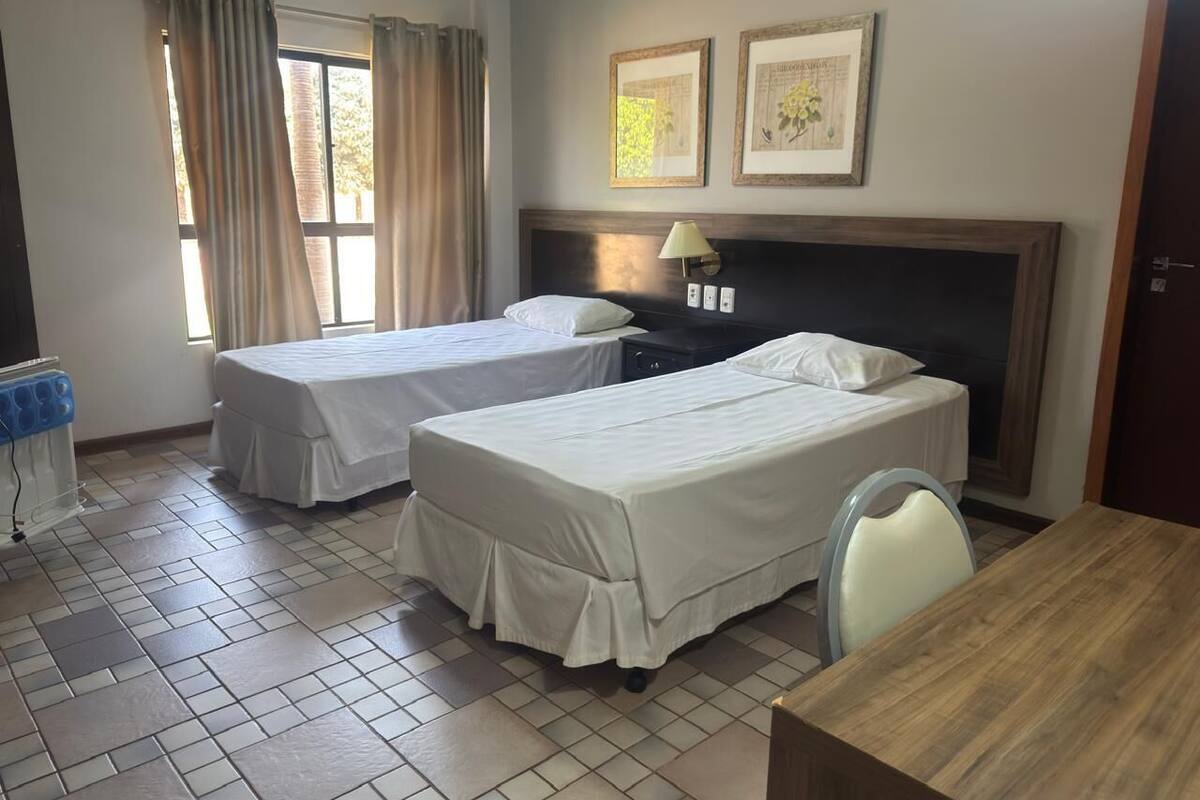 A hotel room with two beds, a wooden table, a chair, a nightstand, a lamp, and a window with curtains.