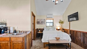 Une chambre avec un grand lit, une table de chevet et un petit tabouret. Il y a également un coin cuisine équipé d’un évier, d’une cuisinière et de placards.