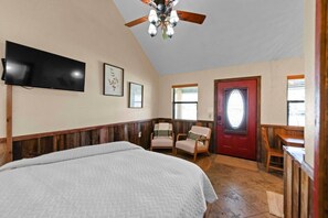 A bedroom with a bed, a TV, a ceiling fan, and a door with a window. - The Tui at Nicole Creek Cabins (Waco)