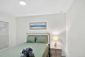Un dormitorio con cama, mesita de noche con lámpara, un cuadro en la pared y ventana con persianas.