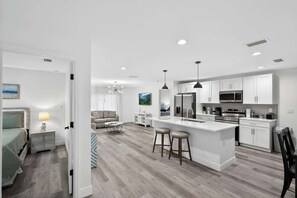 Cocina moderna con armarios blancos, una isla central con taburetes y un comedor con sofá y mesa de centro.