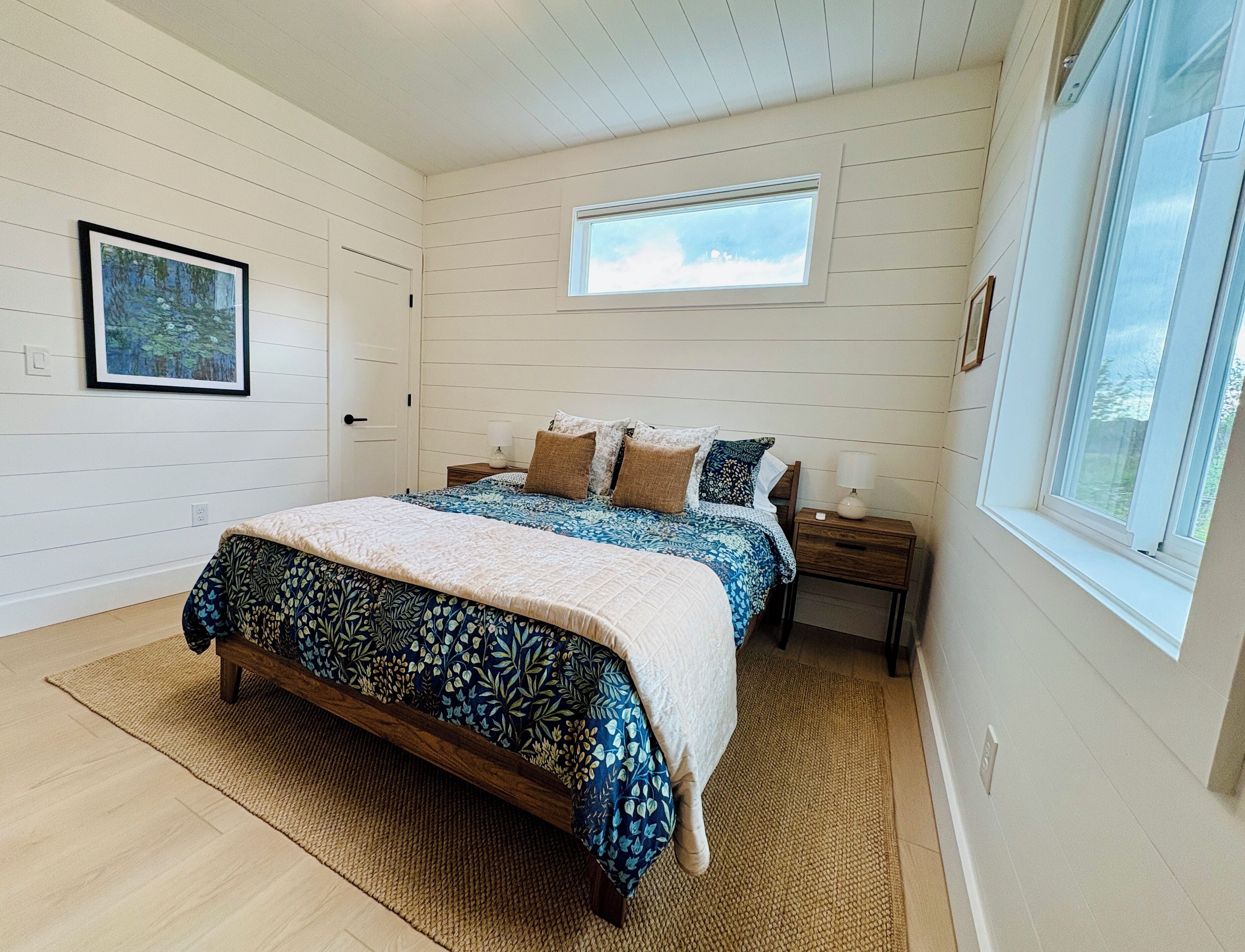 Un dormitorio con cama, mesita de noche, ventana y un cuadro en la pared.