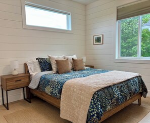 A bedroom with a large bed, a nightstand, a window with a view of trees, and a small picture on the wall.
