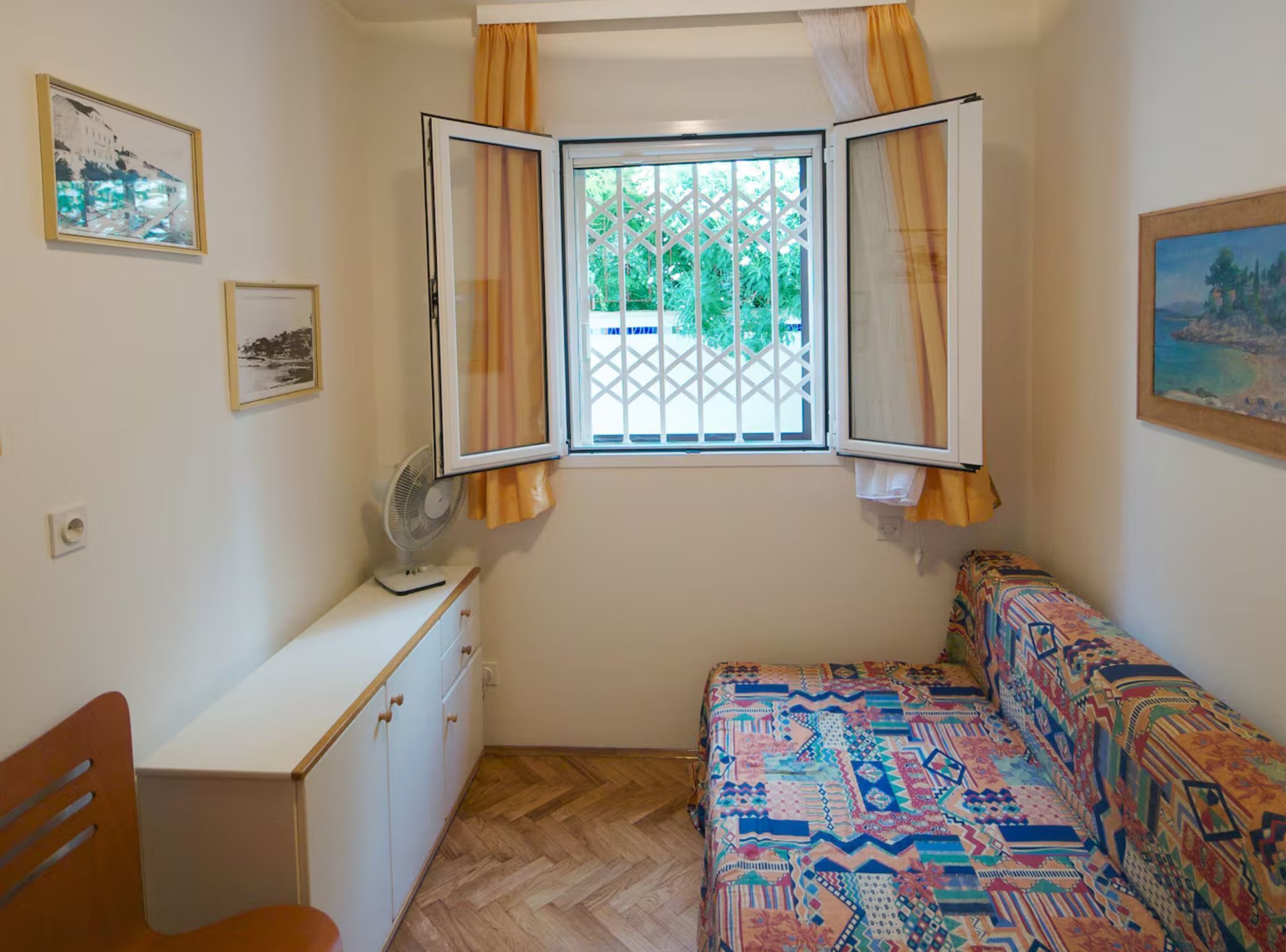 A bedroom with a bed, a desk, a chair, a window with curtains, and two framed pictures on the wall.