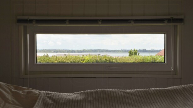 A bedroom with a large window, a bed, and a view of a lake and trees. - Summer house with sea view at Grønsund (Bogø By)