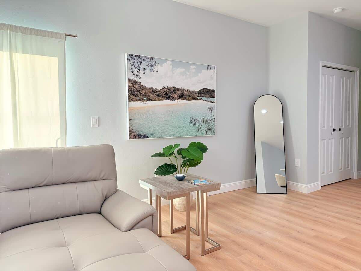 A living room with a sofa, a small table with a plant, a painting of a beach, and a full-length mirror.