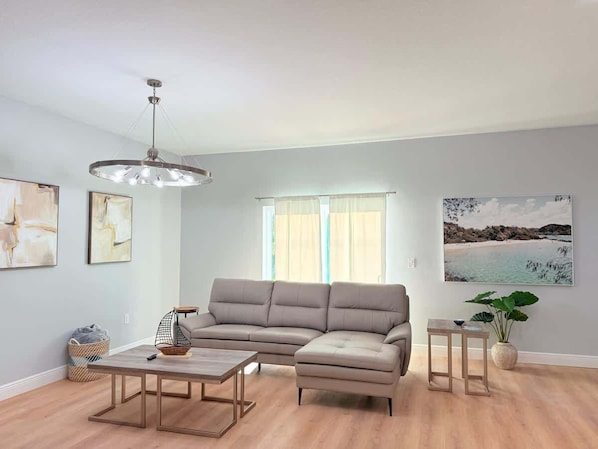 A living room with a sectional sofa, a coffee table, a pendant light, and a large window with sheer curtains.