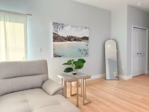 Ein modernes Wohnzimmer mit einem beigen Sofa, einem kleinen Holztisch mit einer Pflanze, einem großen Gemälde zum Strandthema und einem großen Spiegel.