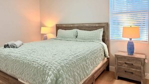 A bedroom with a bed, bedside table, lamp, and a window with blinds. - Bright & Spacious 4/2 Home | Near Beaches & Dining (Port Charlotte)