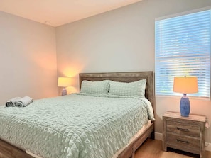 A bedroom with a bed, bedside table, lamp, and a window with blinds.