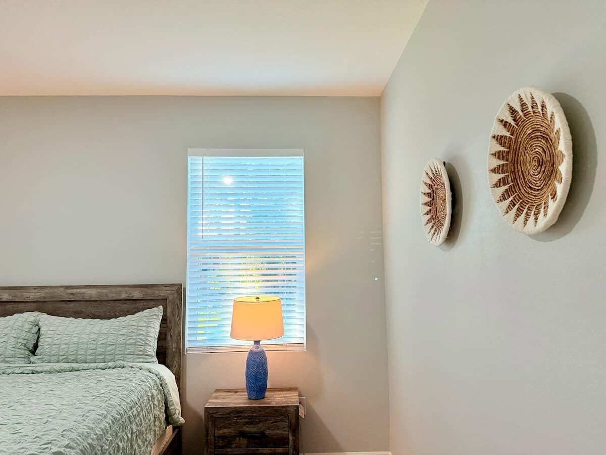 A bedroom with a bed, a window with blinds, a lamp on a nightstand, and two wall decorations.