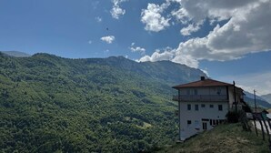 Un edificio blanco con balcón, rodeado de montañas verdes exuberantes y un cielo despejado.