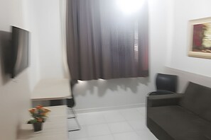 A room with a grey sofa, a small table with a vase of flowers, and a large window with a curtain. - HOTEL BOULEVARD SUITES HOTEL  (Caldas Novas)