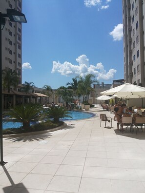 A pool area with tiled flooring, palm trees, and outdoor seating with umbrellas.