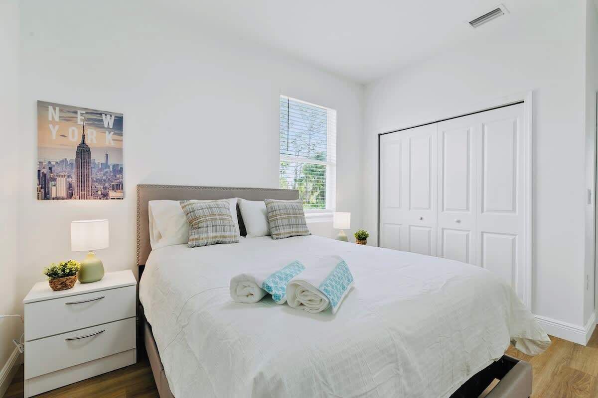 A neatly made bed with rolled towels, a white nightstand with drawers, a lamp, and a framed picture of a cityscape.