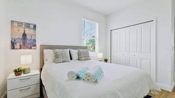 A neatly made bed with rolled towels, a white nightstand with drawers, a lamp, and a framed picture of a cityscape.