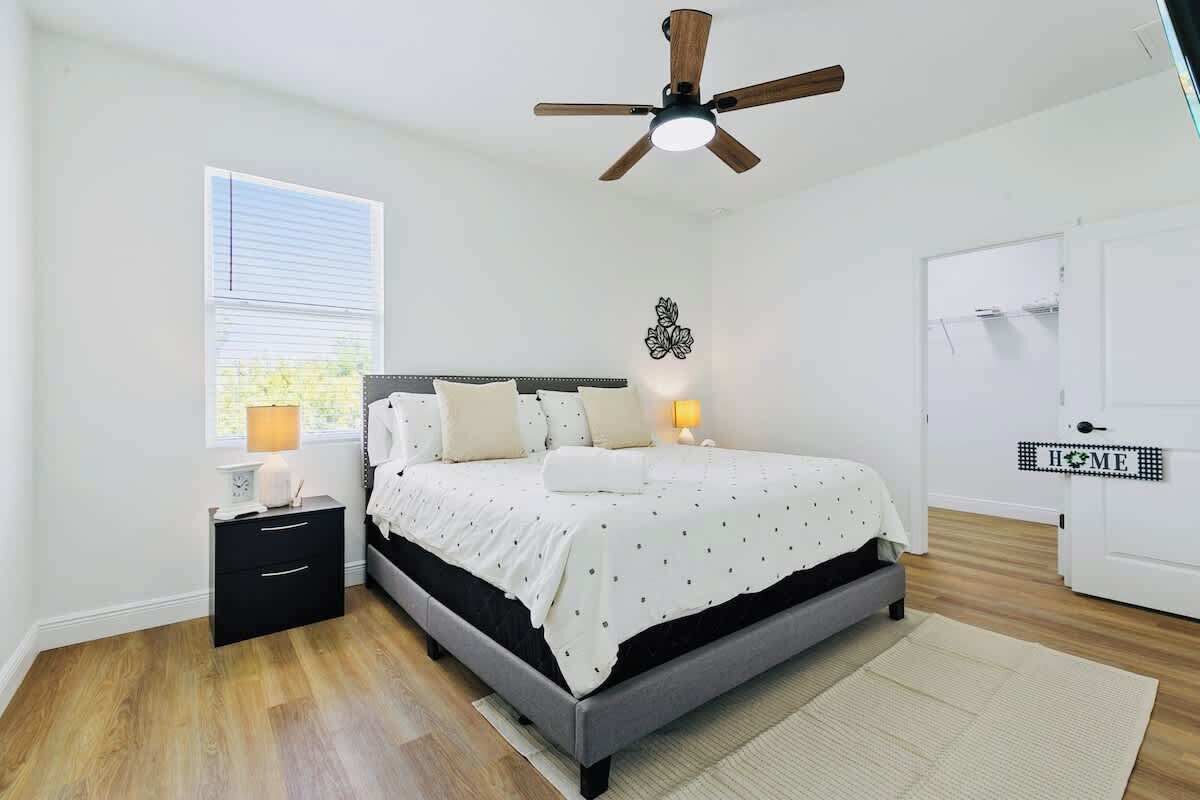 Un dormitorio con cama, mesita de noche, lámpara, ventilador de techo, ventana y una puerta con un letrero que dice 'HOME'.