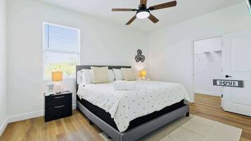 Un dormitorio con cama, mesita de noche, lámpara, ventilador de techo, ventana y una puerta con un letrero que dice 'HOME'.