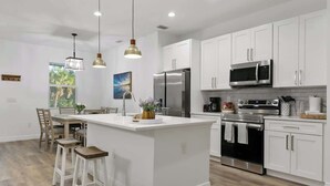 A modern kitchen with white cabinetry, stainless steel appliances, and a central island with bar stools. - Luxe Englewood Home Near Beach (Englewood)