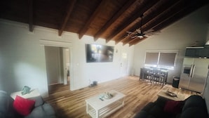 Ein großzügiger Wohnbereich mit Holzboden, einem an der Wand montierten Flachbildfernseher, einer Essecke mit Tisch und Stühlen sowie einer Küchenzeile mit Kühlschrank.