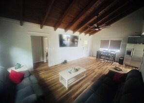 Ein großzügiger Wohnbereich mit Holzboden, einem an der Wand montierten Flachbildfernseher, einer Essecke mit Tisch und Stühlen sowie einer Küchenzeile mit Kühlschrank.