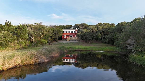 Ein zweistöckiges Haus mit roter Fassade und einer Holzterrasse, umgeben von dichtem Grün und einem ruhigen Teich.